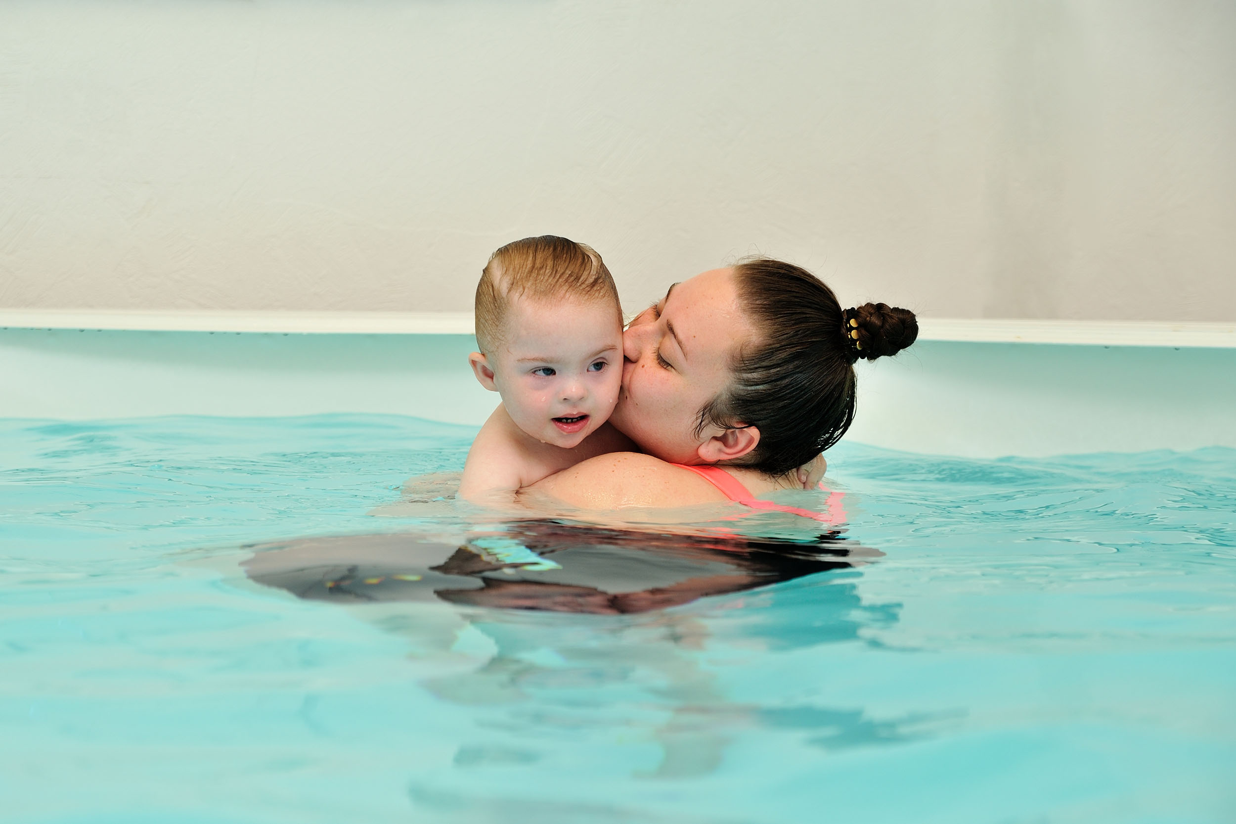 A mother and her child with Down syndrome swim and play in a children’s pool with blue water.